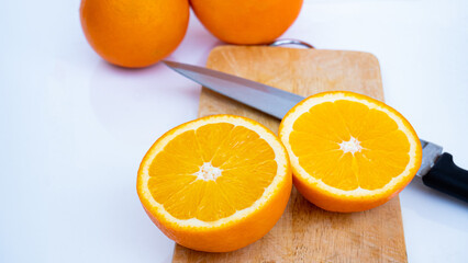Ripe oranges are cut in half and placed on a wooden cutting board. With a sharp knife placed on the ground and a white background