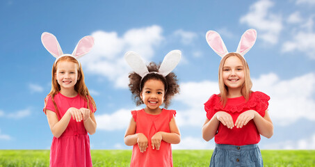 easter, holidays and childhood concept - group of happy smiling girls wearing bunny ears headbands over blue sky and grass background