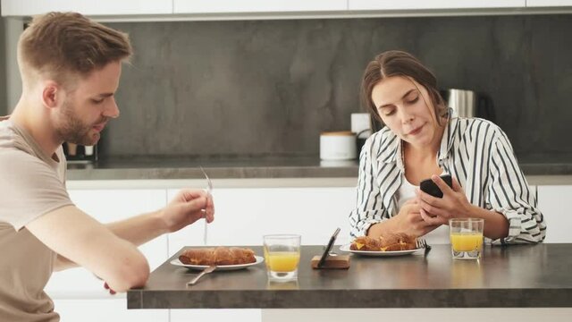 A beautiful young couple man and woman are sitting and using smartphones during breakfast in the kitchen at home