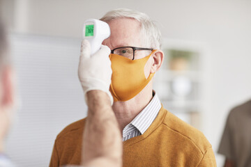 Shot of unrecognizable doctor checking temperature of senior man wearing mask in line at clinic or hospital, copy space