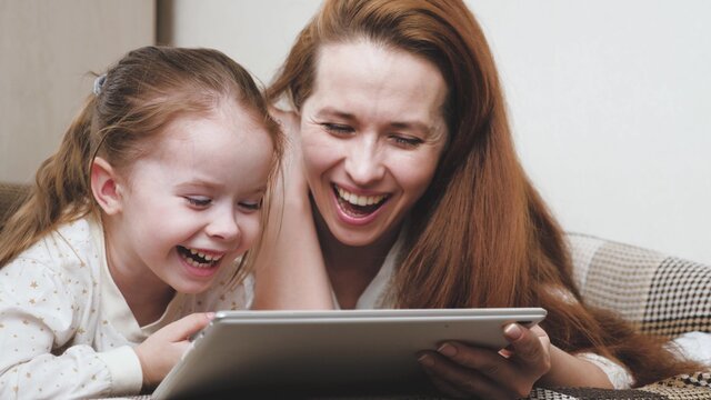 A happy family, mommy and a child are looking at an interesting cartoon on the tablet display and laughing, life is on online learning in the application, mom and daughter are lying on the couch and