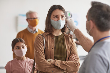 Fototapeta na wymiar Shot of unrecognizable doctor checking temperature of young woman wearing mask and waiting in line at clinic or hospital, copy space