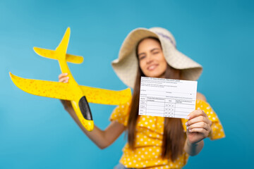 A blurred photo of a young girl in a summer hat holding a illuminating plane in her hand and showing a vaccination card at the camera on her outstretched hand. Blue background.