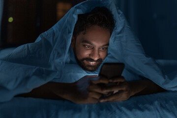 technology, internet, communication and people concept - young indian man with smartphone lying in bed under blanket at home at night