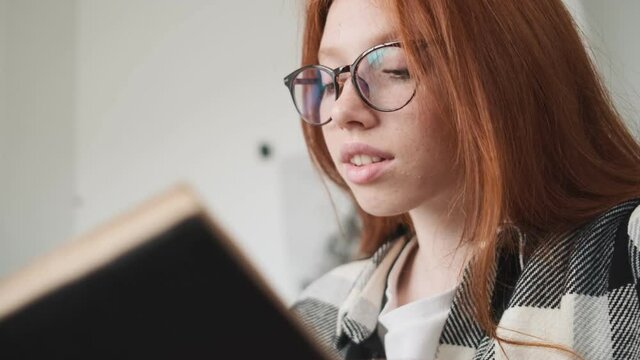 A beautiful redhead woman wearing glasses is reading a book sitting in the apartments