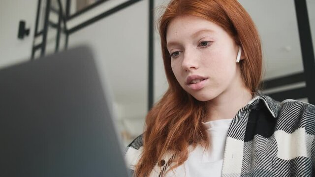 A close-up view of a focused redhead woman with earbuds is using her laptop computer sitting in the apartments