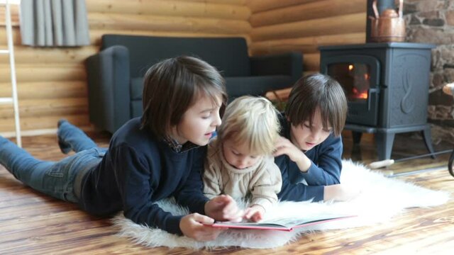 Three sweet children, siblings lying on the floor in little fancy wooden cottage, reading a book, drinking teaand enjoying cozy atmosphere