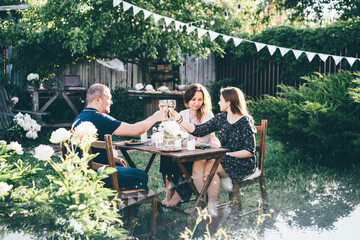 Happy family raise glasses of tasty white wine sitting at small table decorated with peony flowers near wooden shed in green summertime backyard.