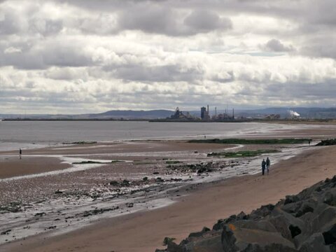 People walk along a dirty looking beach with sea fall defense and industrial landscape in the background