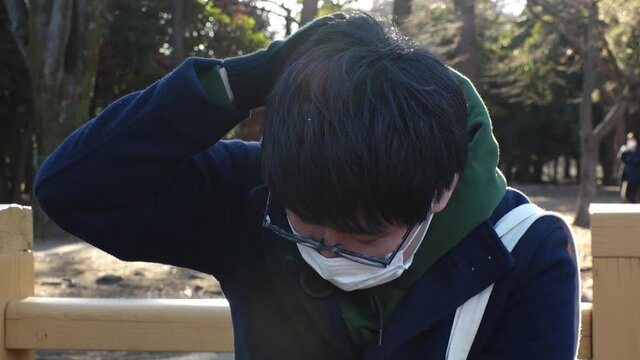 Asian (Japanese) young man with white surgical mask at the park in daytime. Disappointed sad face, one person, late twenties, black hair with glasses. Coronavirus (COVID-19) concept shot. Slow motion.