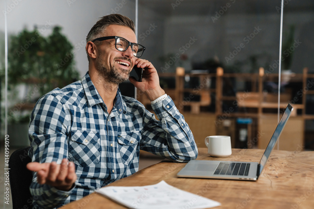 Wall mural smiling grey man talking on mobile phone while working with laptop