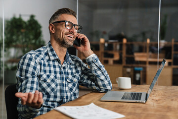 Smiling grey man talking on mobile phone while working with laptop