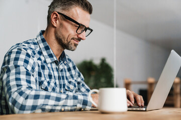 Pleased unshaven man in eyeglasses working with laptop in office