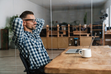 Pleased unshaven man in eyeglasses resting while working with laptop