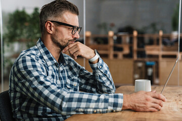 Focused unshaven man working with laptop at table in office