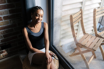 Happy african american girl in earphones using cellphone while sitting at window