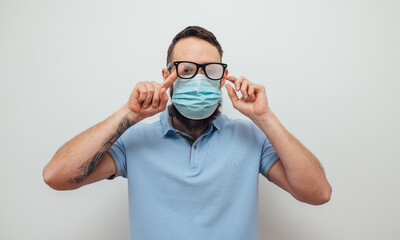 Handsome man with beard wiping blurred foggy misted glasses caused by wearing medical mask on light background. Covid-19