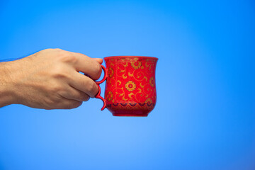 Gold decorated red ceramic cup held in hand by Caucasian male hand isolated on blue background...