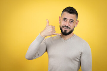 Handsome man with beard wearing sweater over yellow background smiling doing phone gesture with hand and fingers like talking on the telephone