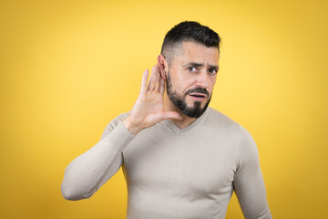 Handsome man with beard wearing sweater over yellow background surprised with hand over ear listening an hearing to rumor or gossip