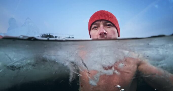 Winter swimmer portrait, cold treatment. Young man soaks in the icy winter lake then turns his head and looks at the camera. Split composition with underwater view
