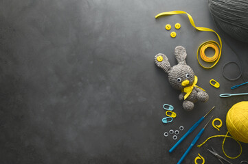 Easter crafting DIY. Handmade knitted toy Easter rabbit, and needlework accessories on dark grey background. Overhead view, space for text, flat lay