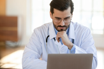 Concentrated young male gp doctor in glasses and medical coat looking at computer screen, thinking...