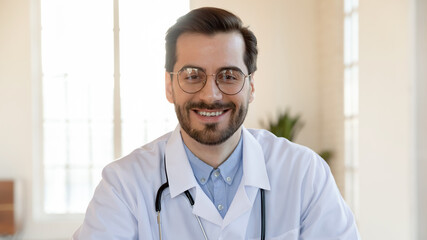 Head shot close up smiling pleasant young handsome family doctor general practitioner in eyeglasses looking at camera, enjoying distant educational webinar or consulting patient online at video call.