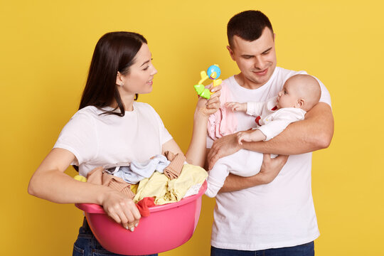 Parents looking after newborn child while doing household chores and laundry, mother with dark hair showing beanbag to their little kid, fatter holds baby in hands, isolated over yellow background.