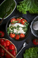 table decorated with fresh mozzarella salad with lettuce and arugula leaves, pesto sauce, fresh cherry tomatoes and cedar nuts, on dark slate table background
