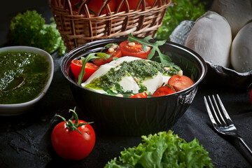 fresh mozzarella salad with lettuce and arugula leaves, pesto sauce, fresh cherry tomatoes and cedar nuts, on dark slate table background
