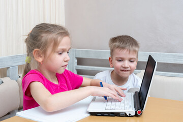Little boy and girl study online at home. Cute little girl student shows how to do homework to her brother. The girl is typing the text on the laptop. Learning concept, distance learning