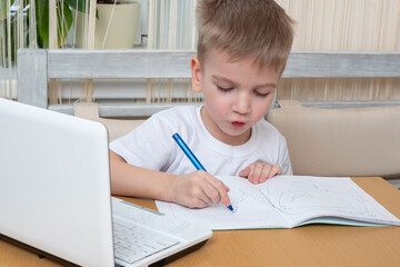 The child studies online at home. Cute little boy draws homework in a notebook sitting at the table in front of a laptop. Online home education concept