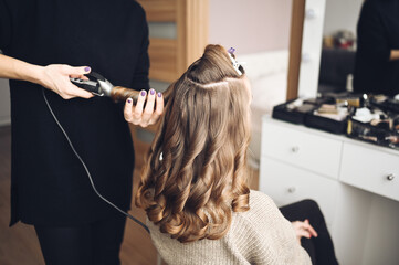 Naklejka premium Hair stylist prepares beautiful young woman for event, makes curls hairstyle with a curling iron for client in beauty salon. Long beautiful light brown natural hair.