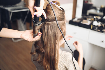 Hair stylist prepares beautiful young woman for event, makes curls hairstyle with a curling iron...