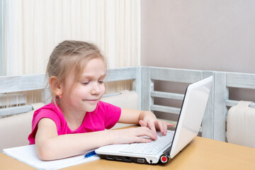 A cute little girl is sitting at a table and typing homework on a laptop at home. Learning concept,...