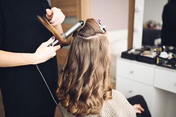 Hair stylist prepares beautiful young woman for event, makes curls hairstyle with a curling iron for client in beauty salon. Long beautiful light brown natural hair.