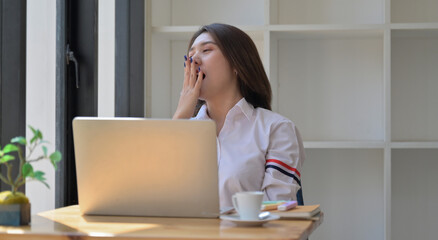 Young woman with a laptop and coffee in the office. She covered mouth with hand and yawned while resting.