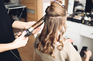 Hair stylist prepares beautiful young woman for event, makes curls hairstyle with a curling iron...