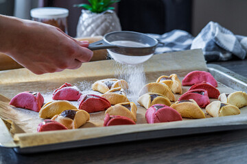 Hand sprinkles sugar powder over colorful hamantaschen cookies in a baking tray. Jewish traditional pastry for Purim (Jewish carnival holiday). Homemade gluten free cookie filled with chocolate.