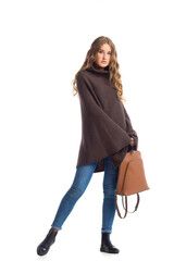 Fototapeta premium Full length portrait of beautiful stylish girl in fashion blue jeans and poncho with long curly hair posing - isolated on white background. Fashion model posing at studio. leather backpack in hand