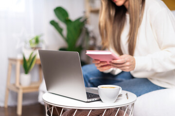 Fototapeta premium Distance learning online education and work. Business woman with coffee mug writes in notebook. Freelance girl with laptop working at home office. Using computer and online shops. Focus on cup