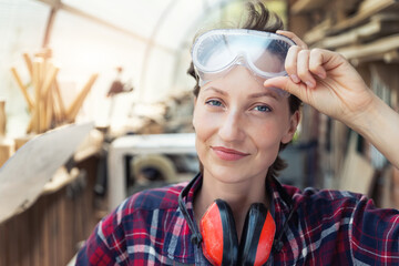 Young beautiful handy professional happy female strong carpenter portrait wearing protective goggles working in carpentry diy workshop against wood. Confident engineer. Women male hobby at workbench