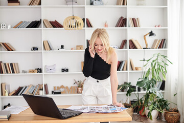 Young blond woman, wearing black top and white pants, standing by table, talking on phone. Office manager at her workplace, preparing for presentation. Female working in light office in company.