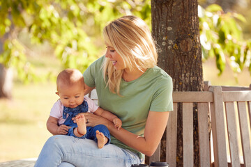 Loving Mother Cuddling And Playing With Baby Daughter Outdoors Sitting On Seat Under Summer Tree