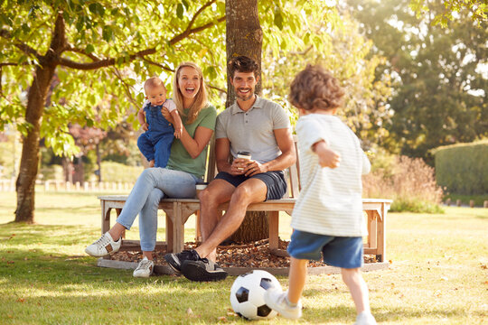 Family With Baby Girl Having Fun In Park Playing Football And Sitting On Seat Under Tree
