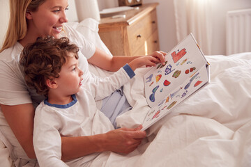 Mother In Bedroom Looking At Picture Book With Young Son Wearing Pyjamas