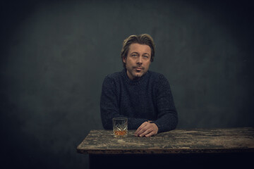 Man in blue sweater with glass of whiskey behind old worn out wooden table.