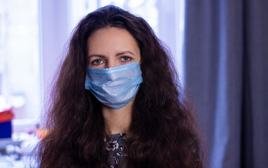 a woman with dark, curly hair wearing a blue, medical mask. Close-up portrait on a gray background