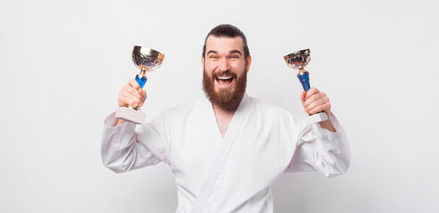 Cheerful smiling man with beard wearing taekwondo uniform and celebrating with two golden cups.
